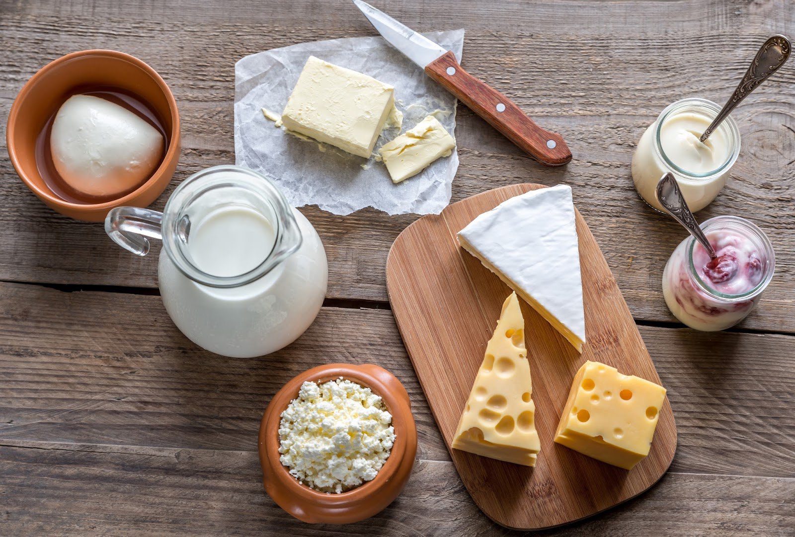 a jar with milk, butter, a wooden board with different kinds of cheese, yogurt, cottage cheese on the wooden table